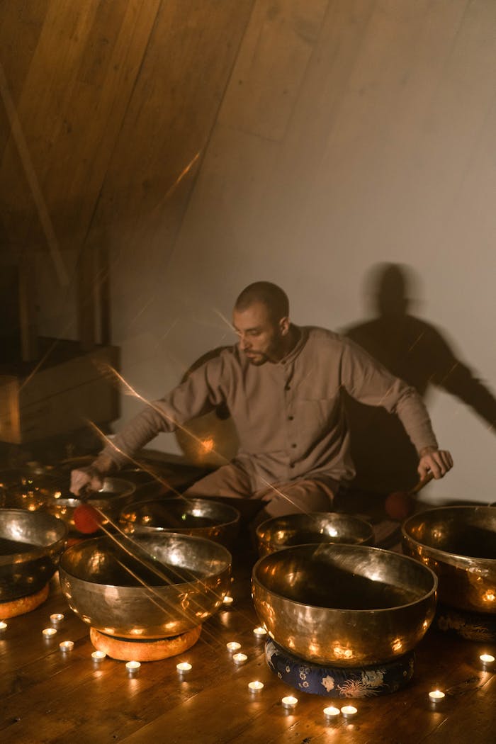 A meditative scene with a man playing Tibetan singing bowls surrounded by candles indoors.